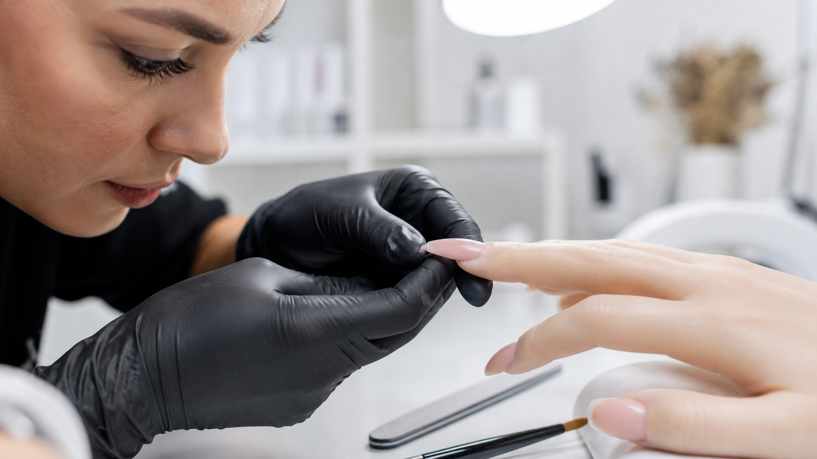 Onicotecnica con guanti neri controlla di profilo l’apex di una ricostruzione unghie nude in gel