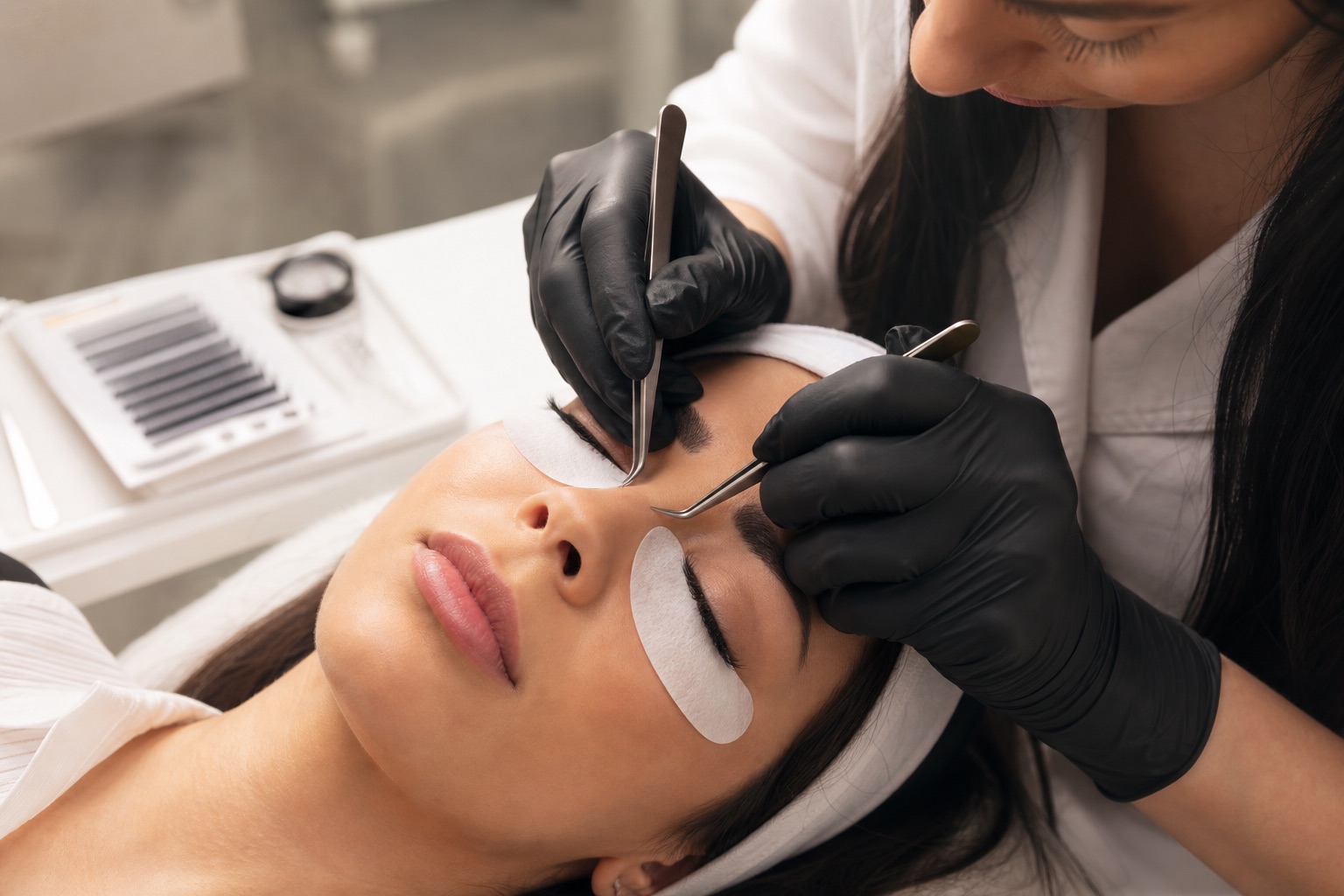 Lash artist adulta applica extension volume a una cliente adulta con occhi chiusi, pad protettivi, guanti, due pinzette e tray di fibre sottili