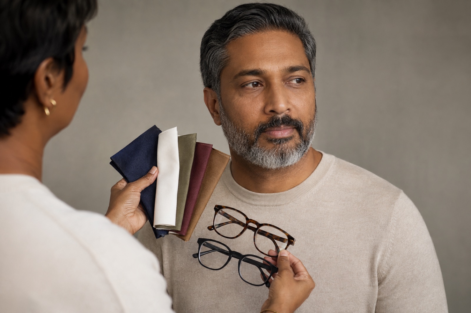 Image consultant compares navy soft white olive burgundy and camel fabrics plus two eyeglass frames beside an adult man with medium-deep skin and a salt-and-pepper beard