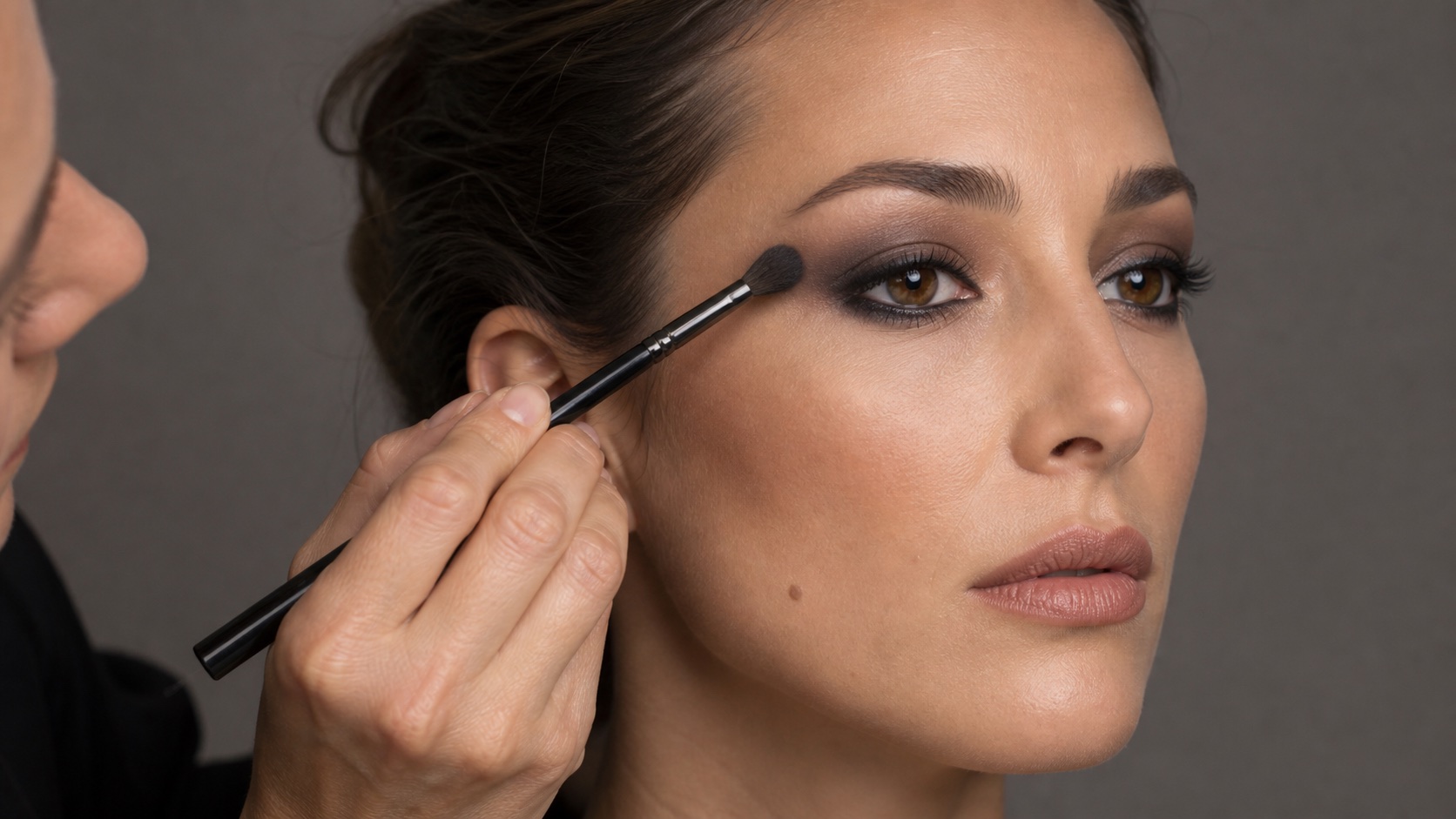 Make-up artist adulta sfuma uno smokey eyes marrone e antracite con un pennello pulito sull’occhio aperto di una modella adulta