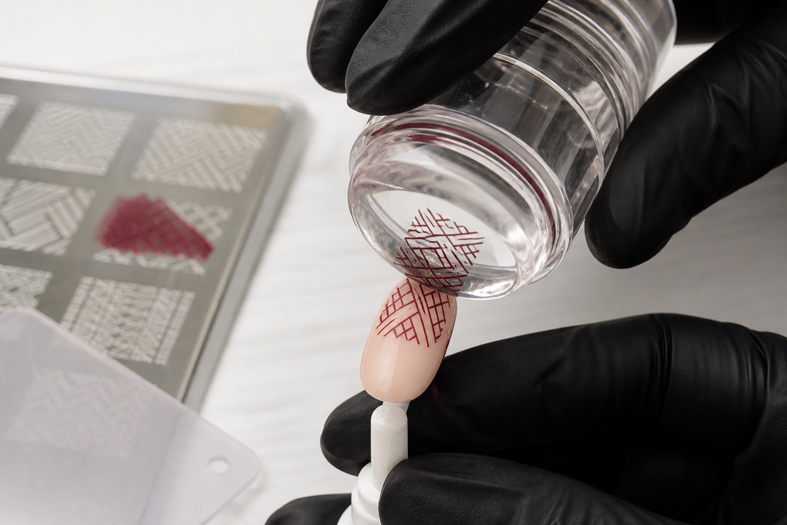Stamper trasparente trasferisce un motivo geometrico burgundy su una tip nude mentre piastra e scraper restano sul piano di lavoro